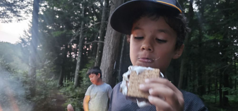 Jasper eating a s'more by the fire, marshmallow on his face, a friend in the background.