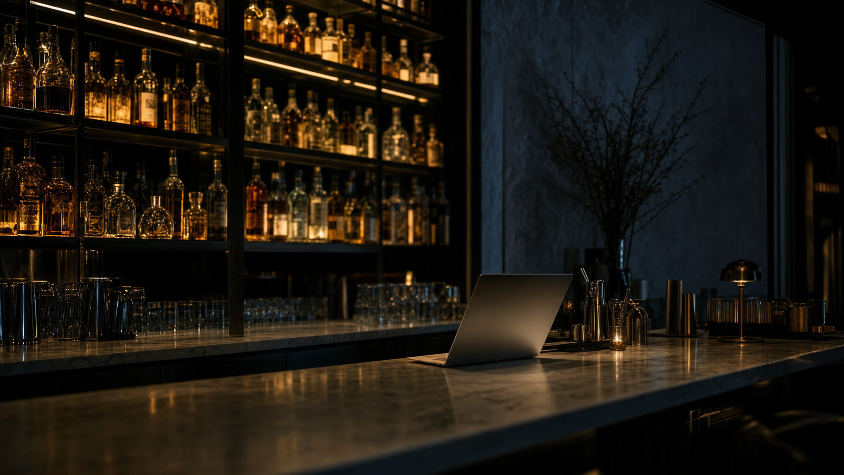 Dark editorial photograph of a NYC spirits back-bar at night with rows of amber bottles and a laptop on a marble bar top.